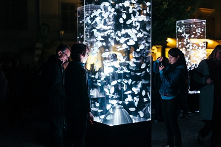 Zwei Männer stehen vor einer Glasvitrine, in der Federn durch ein Gebläse aufgewirbelt werden. Eine Frau macht ein Foto. Aufgenommen beim "Klanglicht" im Rahmen des Designmonats in Graz